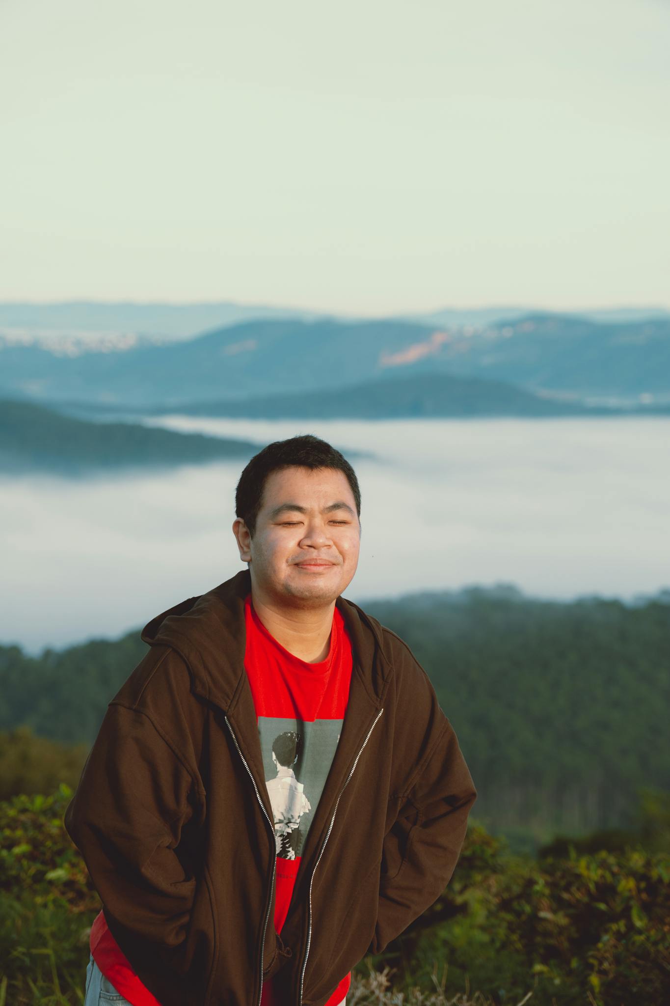 Serene portrait of an Asian man standing against Dalat's misty mountain backdrop, eyes closed, in casual wear.